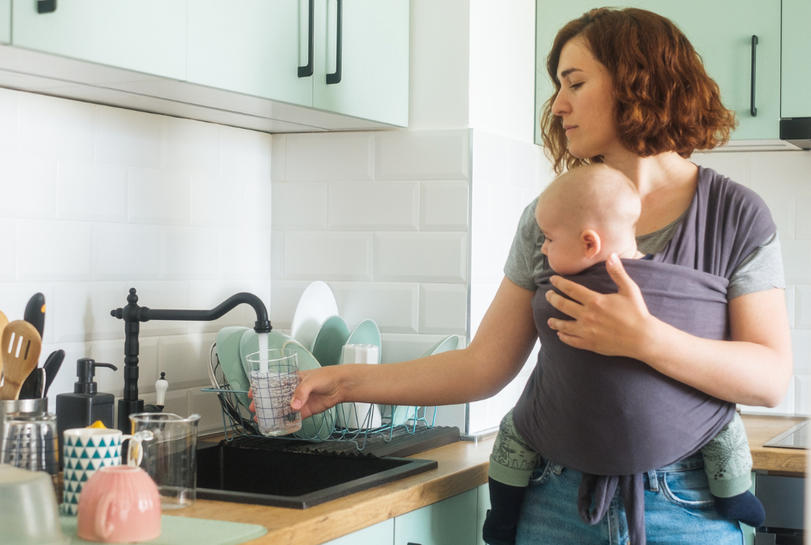 Eine Frau traegt ihr Baby in einem Tragetuch vor dem Bauch und befuellt ein Glas Wasser am Wasserhahn mit einer Hand Eine Frau traegt ihr Baby in einem Tragetuch vor dem Bauch und befuellt ein Glas Wasser am Wasserhahn mit einer Hand