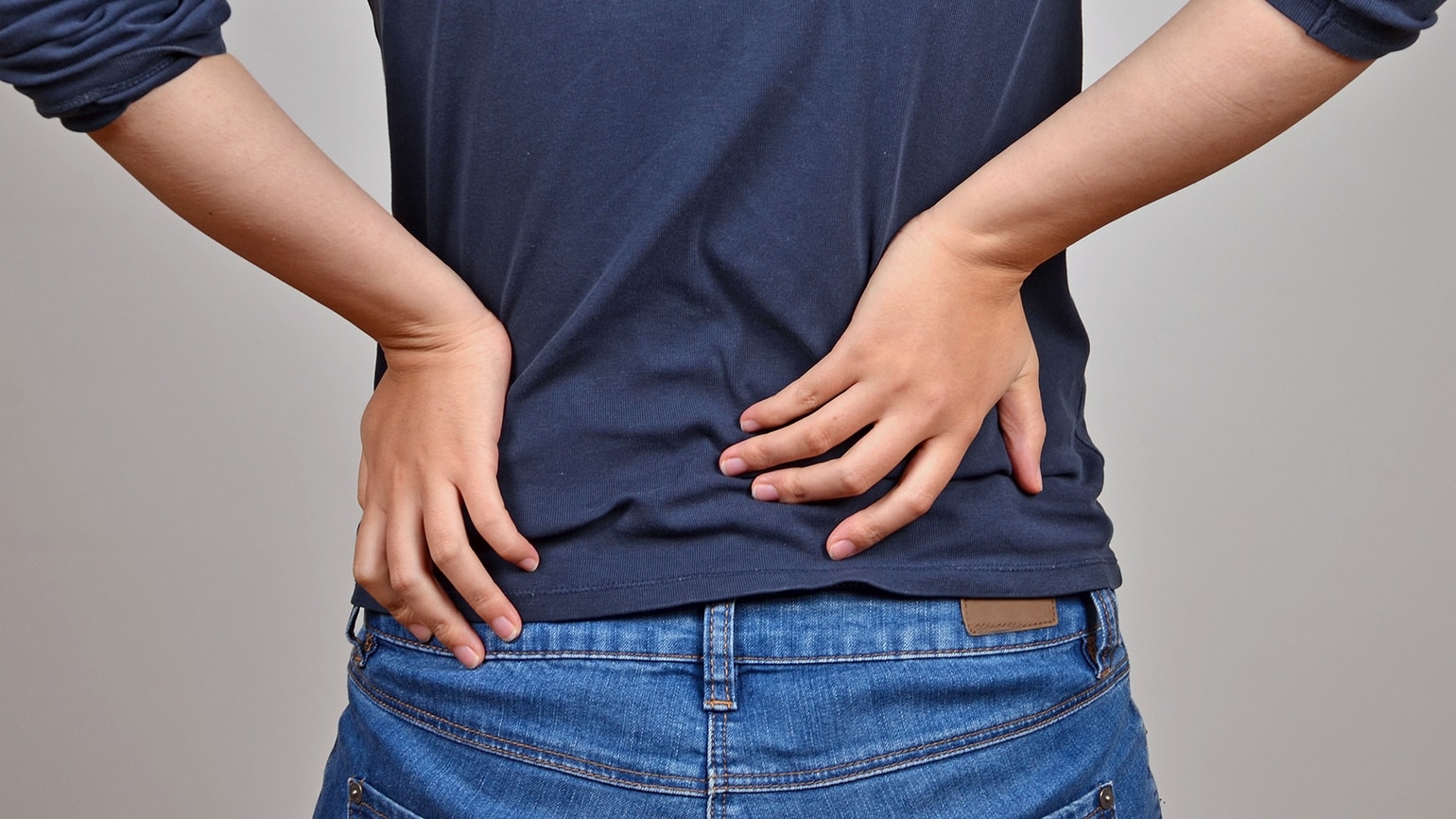 Eine Person in Jeans und T-Shirt hält sich vor Schmerzen den Rücken mit beiden Händen Eine Person in Jeans und T-Shirt hält sich vor Schmerzen den Rücken mit beiden Händen