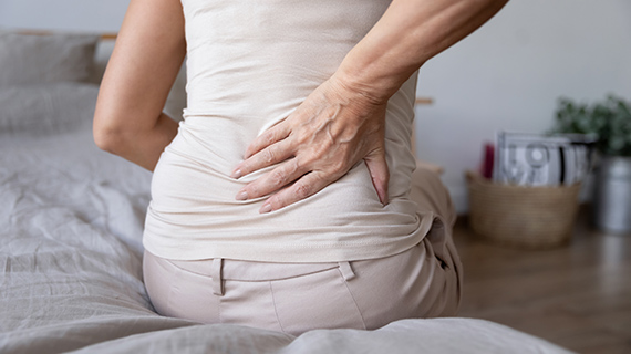 Eine ältere Frau sitzt auf dem Bett und hält sich die rechte Hand an den unteren Rücken Eine ältere Frau sitzt auf dem Bett und hält sich die rechte Hand an den unteren Rücken
