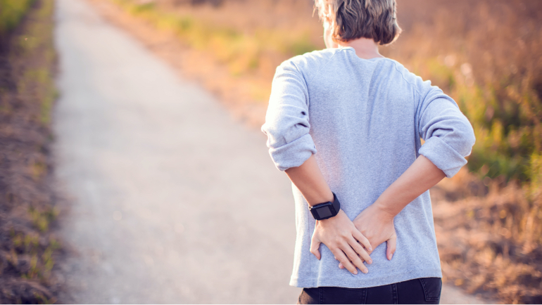 Eine Person geht auf einem Feldweg und haelt sich mit beiden Haenden den unteren Ruecken Eine Person geht auf einem Feldweg und haelt sich mit beiden Haenden den unteren Ruecken