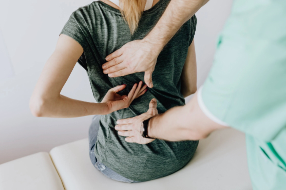 Frau sitzt auf einer Liege sie fasst sich mit einer Hand in den Ruecken ein Mann steht hinter ihr und sie mit beiden Haenden an Frau sitzt auf einer Liege sie fasst sich mit einer Hand in den Ruecken ein Mann steht hinter ihr und sie mit beiden Haenden an