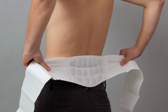 Mann mit freiem Oberkörper legt ThermaCare® Wärmeumschlag gegen Rückenschmerzen von hinten um Mann mit freiem Oberkörper legt ThermaCare® Wärmeumschlag gegen Rückenschmerzen von hinten um